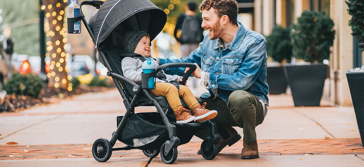 An adult kneeling beside a child in a Chicco lightweight stroller on a sidewalk